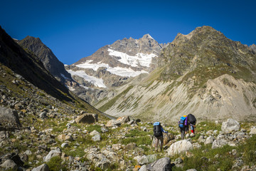 Fototapeta premium Group of Hikers male and female walking on Mountain Trail. carrying backpacks using trekking sticks rear. mixed grassy and rocky terrain. View People are unrecognizable behind