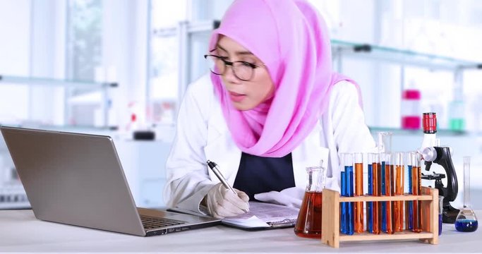 Muslim scientist making research report on the clipboard while looking at the laptop with test tube on the table in the lab, shot in 4k resolution
