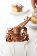 
Chocolate frosting in small white bowl with a slice of yellow cake on white background with spreading knife
