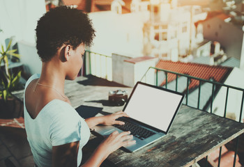 Rear view of young curly Brazilian female working on the balcony with her laptop with white screen...