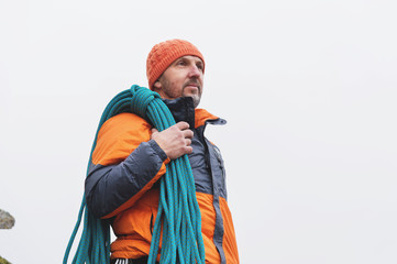 Hipster-climber stands in a down jacket on a rock with a rope on his shoulder