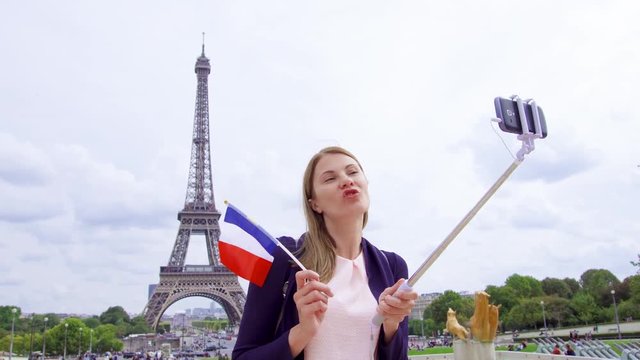 Woman with small French flag near Eiffel Tower in Paris, France doing selfie on mobile phone with selfie-stick. Happy smiling tourist woman traveling in Europe.