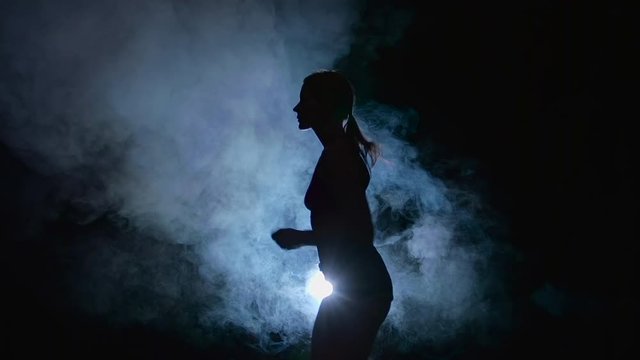 Athletic fitness girl running on a black background illuminated by the spotlight in the smoke. Silhouette. Slow motion