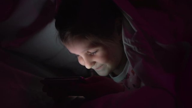 Little cute girl is looking at a smartphone cartoon lying on a bed under a blanket at night.
