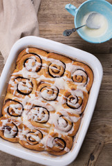 Glazed cinnamon buns  in a baking dish. Frosting on the top right corner. Wooden tabletop 