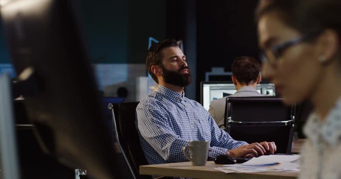 Handsome man with a beard working late at evening on the computer surrounded by others employees working at their computers. Businessman work. Inside nice office room
