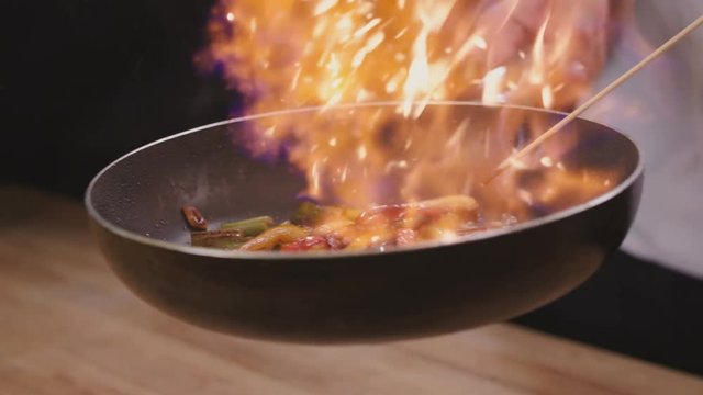 Close up of a chef sauteing vegetables and sausage in a pan in a kitchen, with flames