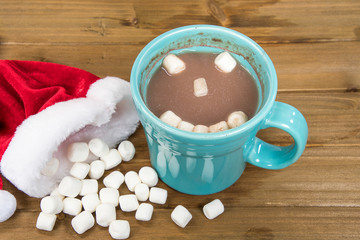 Christmas hot chocolate drink in turquoise mug with Santa cap and marshmallow on rustic wood