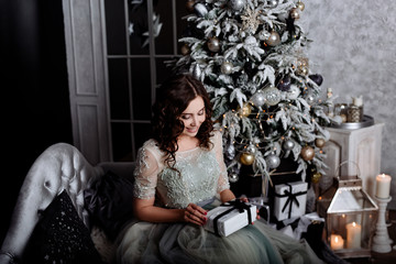 Obraz na płótnie Canvas The young woman the brunette with curly hair in an elegant blue dress against the background of the room which is beautifully decorated by Christmas. The girl smiles and opens a Christmas gift.