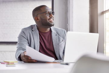 Obraz premium Happy dark skined male manager wears classic suit, works on computer and with documents, sits at spacious cabinet, makes banking online. Cheerful middle aged African male owner of business company