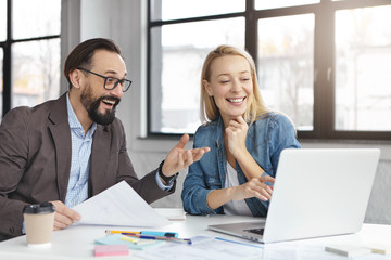 Indoor shot of successful businessman and businesswoman look happily at laptop, glad to see rapid growth of sales, read information about creating online stratup. Great business achievements