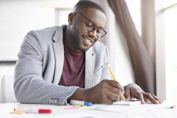 Smiling dark skinned male enterpreneur writes notes in notepad, sits at coworking space. African...