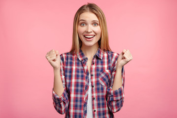 Waist up portrait of delightful smiling happy woman dressed in checkered shirt, clenches fists, rejoices good news, being confident in her big success, isolated over pink background. I have done it!