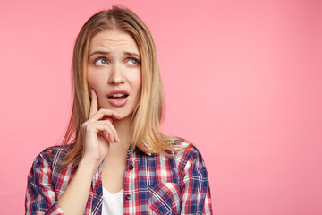 Horizontal portrait of attractive blonde female has unhappy look, thinks how to solve her problem, looks up, poses against pink background with copy space for your promotional text or advertisment