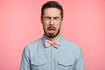 Bearded brunet attractive man closes eyes and opens mouth sees something sour, isolated over pink...