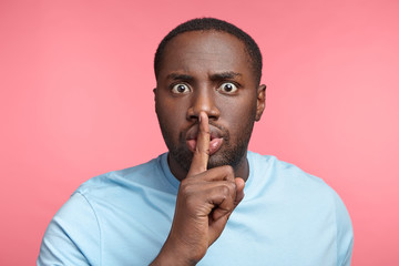 Buged eyed dark skinned male keeps fore finger on lips, being shocked as friends tells very private information, demands keep silence and stop talking. African male demonstrates shushing gesture