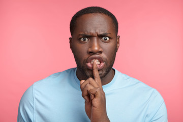 Portrait of angry irritated dark skinned male asks keep confidential information in secret, annoyed with people who gossip, demands quiet, says: Hush, stop talking. African man makes silence gesture