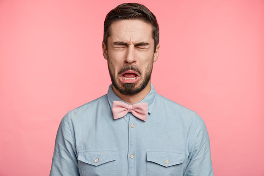 Bearded brunet attractive man closes eyes and opens mouth sees something sour, isolated over pink background. Unhappy handsome young male wears pink bow tie and shirt, makes grimace or pulls face