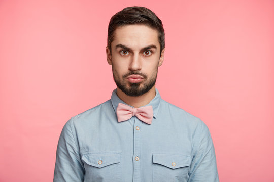 Serious brunet man dressed in formal shirt and bow tie, pouts lips, meets guests who came to congratulate him with anniversary, poses against pink studio background. People and emotions concept