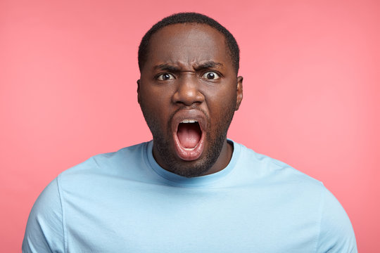 Furious dark skinned male screams loudly, annoyed with interlocutor, expresses negative emotions and feelings, keeps mouth widely opened. Angry African boss shouts at coworkers, being strict