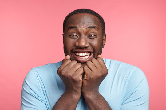 Cheerful emotional happy dark skinned male grins at camera, demonstrates white perfect teeth, keeps fists under chin, glad to recieve positive news that he will become father, isolated over pink wall