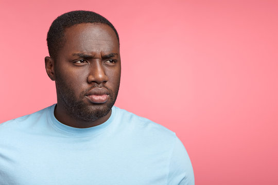 Horizontal portrait of black serious male has full lips, looks aside with thoughtful gloomy expression, feels frustrated and troublesome, poses against pink background with copy space for advertisment