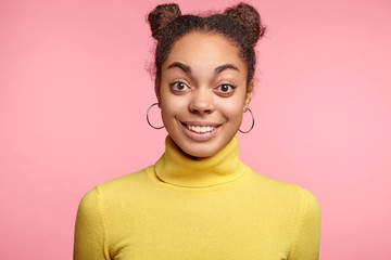 Happy excited surprised female model with two hair buns, raises eyebrows as being amazed with something, can`t believe in her success, glad to be praised, poses against pink studio background
