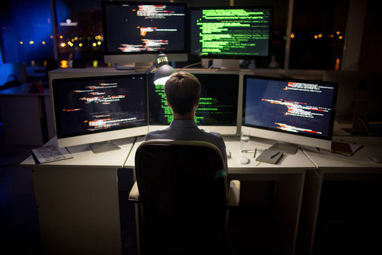 Rear view of hard-working programmer with stylish haircut sitting in front of computer and writing code, interior of dim open plan office on background