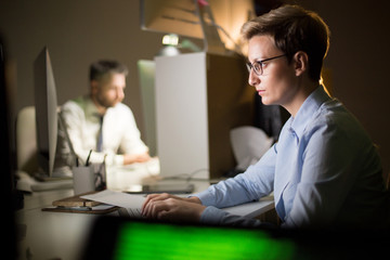 Confident middle-aged manager with stylish haircut working on computer while her male colleague sitting at office desk and analyzing work results, interior of open plan office on background