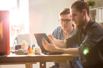 Profile view of bearded young engineer showing work results to his colleague with help of digital tablet while having working meeting at modern laboratory, 3D printer and small prototypes on desk