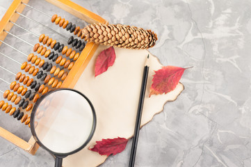 Old wooden abacus and magnifier with black plastic handle and cone and black pencil and three red leaves on burnt paper on cracked gray concrete