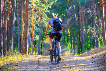Naklejka premium A guy in a gray T-shirt, black shorts and a black backpack is riding along a path in the forest on a bicycle.