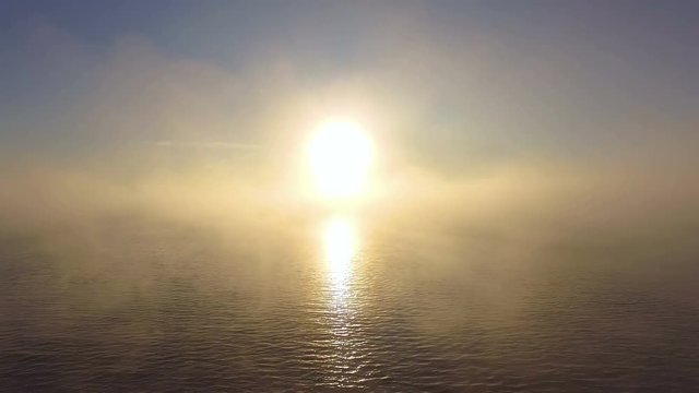 AERIAL: Flight through the mist to bright sun above the water. Sun path on water surface. Appearing coast of the lake.