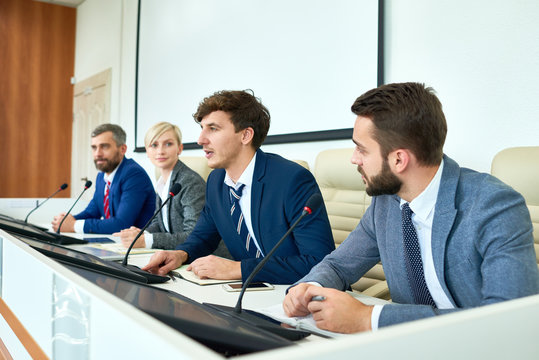 Portrait Of Several Business People Sitting In Row Participating In Political Debate During Press Conference Answering Media Questions Speaking To Microphone