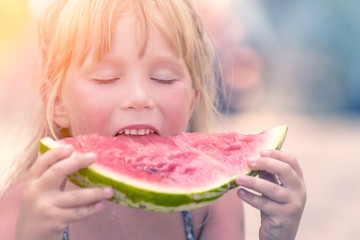 Little cute blond girl eating a slice of red watermelon on blurred background