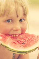 Little cute blond girl eating a slice of red watermelon on blurred background