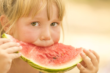 Little cute blond girl eating a slice of red watermelon on blurred background. Close-up, sunny
