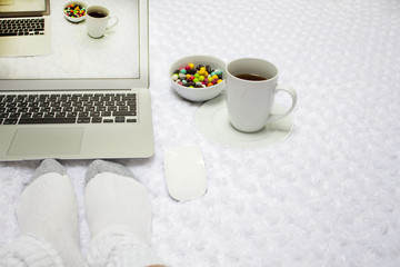 Laptop, a cup of tea, lollipops and female feet in white socks on a white blanket .  the image on the screen comes to life .  reality and picture on the screen are the same. Concept of escape to real.