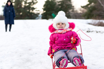 The child, a little girl riding on a sled with snow slides. Winter fun for children. A child with his mother and a winter walk.