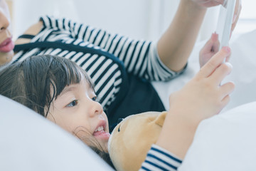 A little cute girl lying with her mother on the white bed in the bedroom, daughter hug her teddy bear and watching cartoon by tablet with her mom, rest time