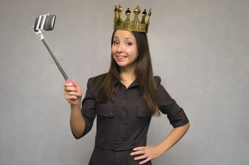 Young caucasian girl with golden crown above her head makes selfie photo on mobile phone by using selfie stick and smiles into camera.