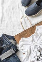 Womens clothing shopping background. Jeans, sneakers, t-shirt, scarf and a paper bag on a light background, top view. Walk stylish look