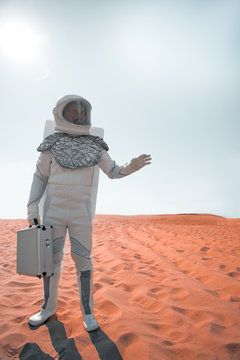 Under hot sun. spaceman is standing at red sand and holding suitcase. He looking aside with seriousness. Full length portrait. Copy space on right side