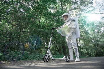 Concentrated spaceman is standing at road in forest and attentively reading unfolded map. He standing near scooter. Low angle. Copy space on left side