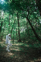 Exploration of new place. Spaceman wearing uniform is standing among trees and holding small suitcase. Profile. Copy space on right side