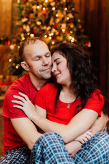 adult married couple in red T-shirts and pajamas embraces sitting on the floor against the Christmas tree.