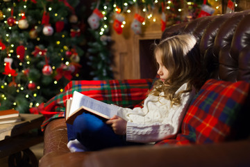 Happy little girl reading a story book by on the couch in a cozy