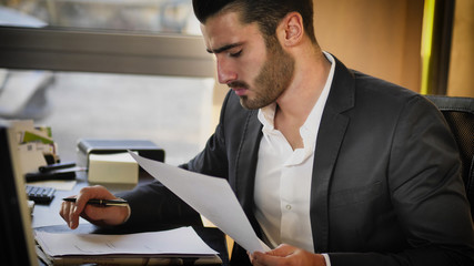 Fototapeta premium Successful handsome young businessman sitting at his desk in the office and looking at the computer screen while writing on sheet of paper
