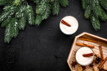 Homemade classic eggnog with cinamon and badian on wooden tray near spruce branch, pinecones on black background top view copyspace