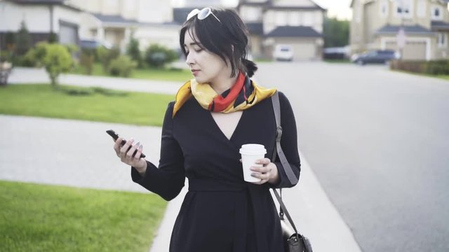 Smiling and beautiful Asian bissinesswoman with a coffee to go is looking at her phone and walking in a suburbian area. Tracking real time establishing shot